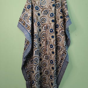 May include: A floor-length kaftan dress featuring a white base with a repeating blue and brown floral pattern. The garment has wide sleeves, a round neckline, and a drawstring waist tie with a striped pattern that matches the sleeve edges.