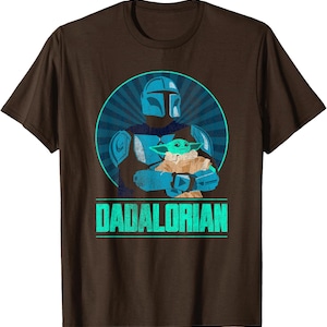 以下が含まれることがあります： 茶色のTシャツ。水色と青色のマンダロリアンがグローグーを抱いているグラフィックが描かれ、青色の放射状の背景が特徴です。「DADALORIAN」の文字が下に水色で表示されています。