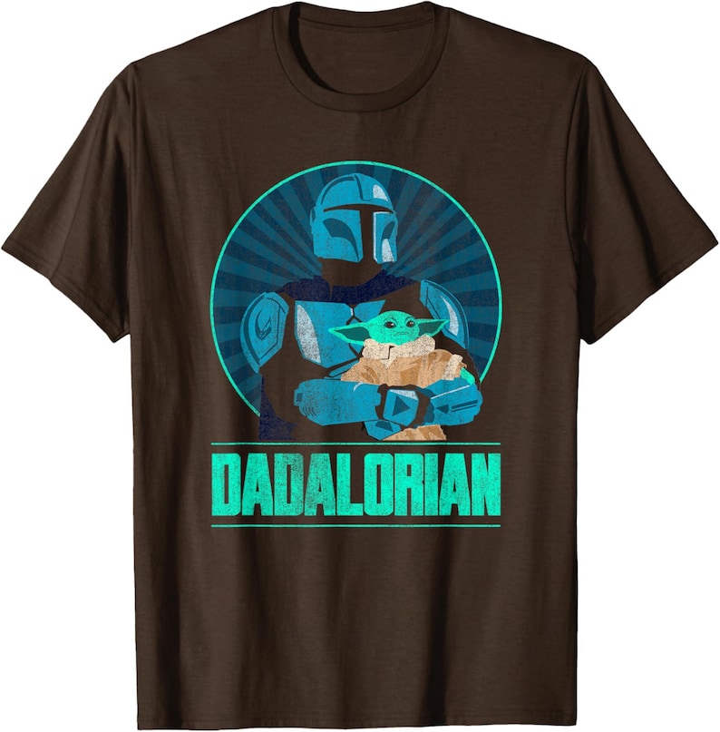 以下が含まれることがあります： 茶色のTシャツ。水色と青色のマンダロリアンがグローグーを抱いているグラフィックが描かれ、青色の放射状の背景が特徴です。「DADALORIAN」の文字が下に水色で表示されています。