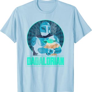 以下が含まれることがあります： 水色のTシャツ。マンダロリアン風のキャラクターが緑色のエイリアンを抱いているグラフィックが描かれています。グラフィックは、使い古された質感の青い円の中に配置されています。「DADALORIAN」の文字が緑色でプリントされています。