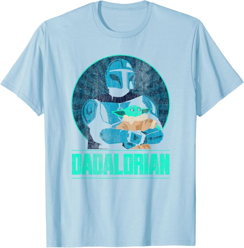 以下が含まれることがあります： 水色のTシャツ。マンダロリアン風のキャラクターが緑色のエイリアンを抱いているグラフィックが描かれています。グラフィックは、使い古された質感の青い円の中に配置されています。「DADALORIAN」の文字が緑色でプリントされています。