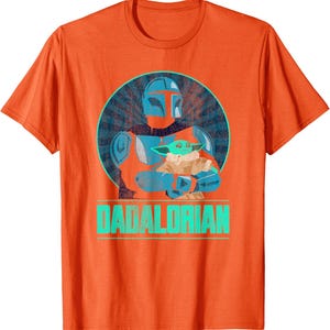 以下が含まれることがあります： オレンジ色のTシャツ。青い円の中に、マンダロリアンキャラクターがグローグーキャラクターを抱いているグラフィック。テキスト「DADALORIAN」はターコイズ色で下に印刷されています。