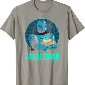 以下が含まれることがあります： グレーのTシャツ。水色と青色のグラフィックで、緑色のエイリアンを抱えるマンダロリアンキャラクターが描かれています。「DADALORIAN」の文字が水色でプリントされています。デザインは、サンバースト効果のある青い円の中にあります。