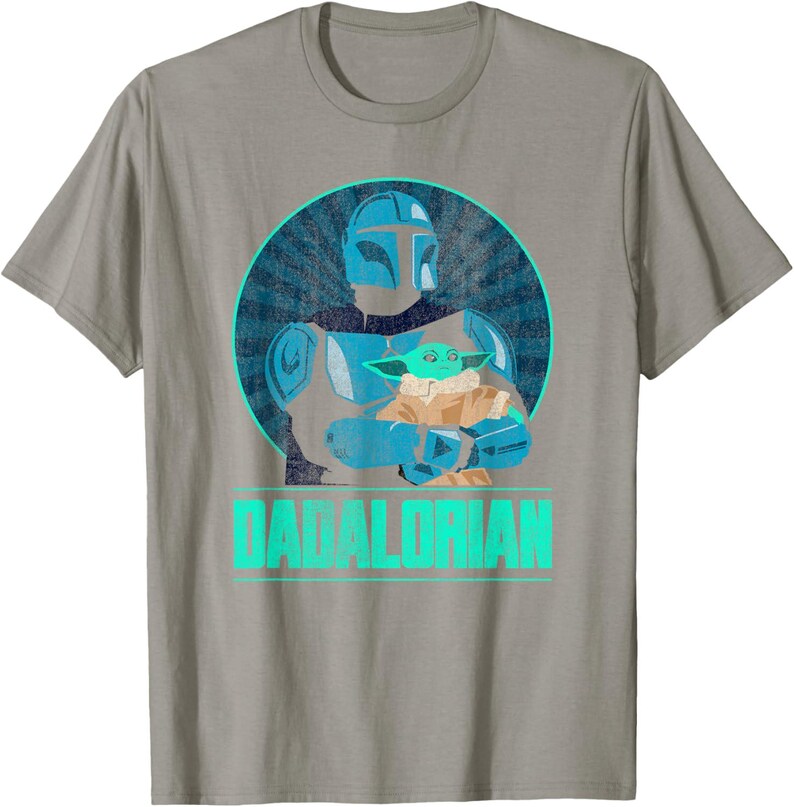 以下が含まれることがあります： グレーのTシャツ。水色と青色のグラフィックで、緑色のエイリアンを抱えるマンダロリアンキャラクターが描かれています。「DADALORIAN」の文字が水色でプリントされています。デザインは、サンバースト効果のある青い円の中にあります。