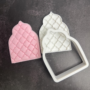 Peut inclure: Trois emporte-pièces en forme de mosquée. L'un est rose, avec un motif texturé, et les deux autres sont blancs. Les emporte-pièces sont en plastique et sont destinés à la pâtisserie. Le fond est une surface gris foncé.