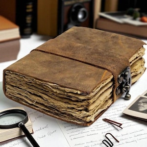 May include: A thick, rectangular journal bound in distressed brown leather with a wrap-around strap and a dark metal latch. The book contains numerous pages of textured, cream-colored paper with deckled edges, giving it a weathered appearance.