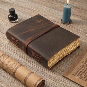 May include: A dark brown leather journal with a wrap-around strap closure and visible cross-stitched binding on the spine. The journal features thick, deckled-edge pages and is positioned near a small black ink bottle and a blue pillar candle.