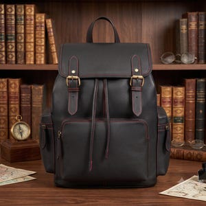 May include: A black leather backpack featuring a top flap with two brass buckle closures, a drawstring main compartment, and two side pockets. The bag is accented with prominent red stitching along the seams and edges, including the straps.