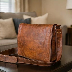 May include: A rectangular brown leather messenger bag with a large front flap, a long adjustable shoulder strap, and visible stitching along the edges. The leather has a smooth, polished finish with natural variations in the tan and brown tones.