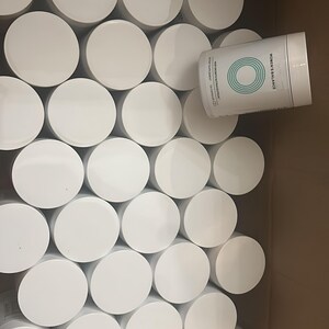 May include: A cardboard box filled with numerous white, cylindrical containers with white lids. One container is partially visible, displaying the text "WOMEN'S BALANCE" and a teal circular design.
