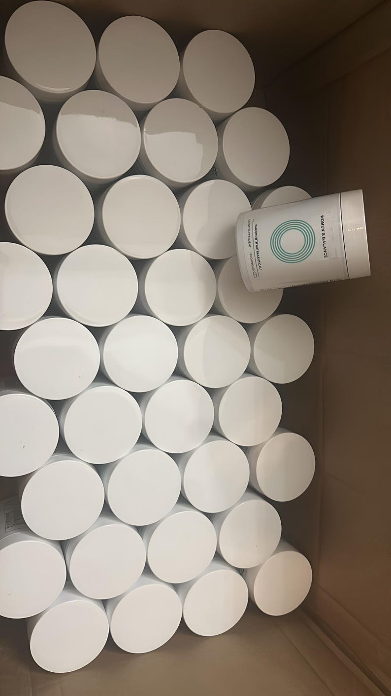 May include: A cardboard box filled with numerous white, cylindrical containers with white lids. One container is partially visible, displaying the text "WOMEN'S BALANCE" and a teal circular design.