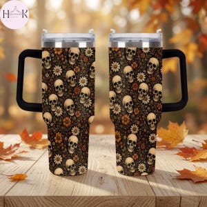 May include: Two stainless steel tumblers with black handles and clear lids, featuring a repeating pattern of beige human skulls interspersed with orange and white flowers on a dark brown background, displayed side by side on a wooden surface.