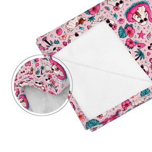 Puede incluir: Una manta suave con un patr&oacute;n repetitivo de fantasmas sobre fondos rosas, acentuado con flores de hibisco tropicales, palmeras, conchas marinas y estrellas de mar. La tela tiene un reverso de felpa blanca y un recuadro circular muestra la textura.