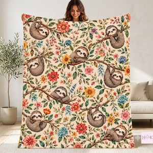 May include: A cream-colored throw blanket featuring a repeating pattern of brown sloths hanging from tree branches, interspersed with colorful red, orange, yellow, and blue flowers and green leaves. The fabric has a soft, plush texture.