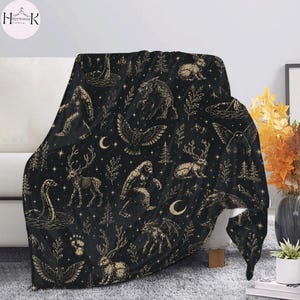 May include: A black throw blanket featuring a repeating gold-toned pattern of mythical creatures including Bigfoot, the Loch Ness monster, jackalopes, and owls, interspersed with crescent moons, stars, pine trees, and various botanical elements.