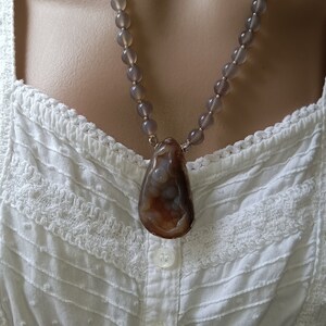 May include: A gray beaded necklace with a large brown agate pendant. The necklace is worn on a white lace top.