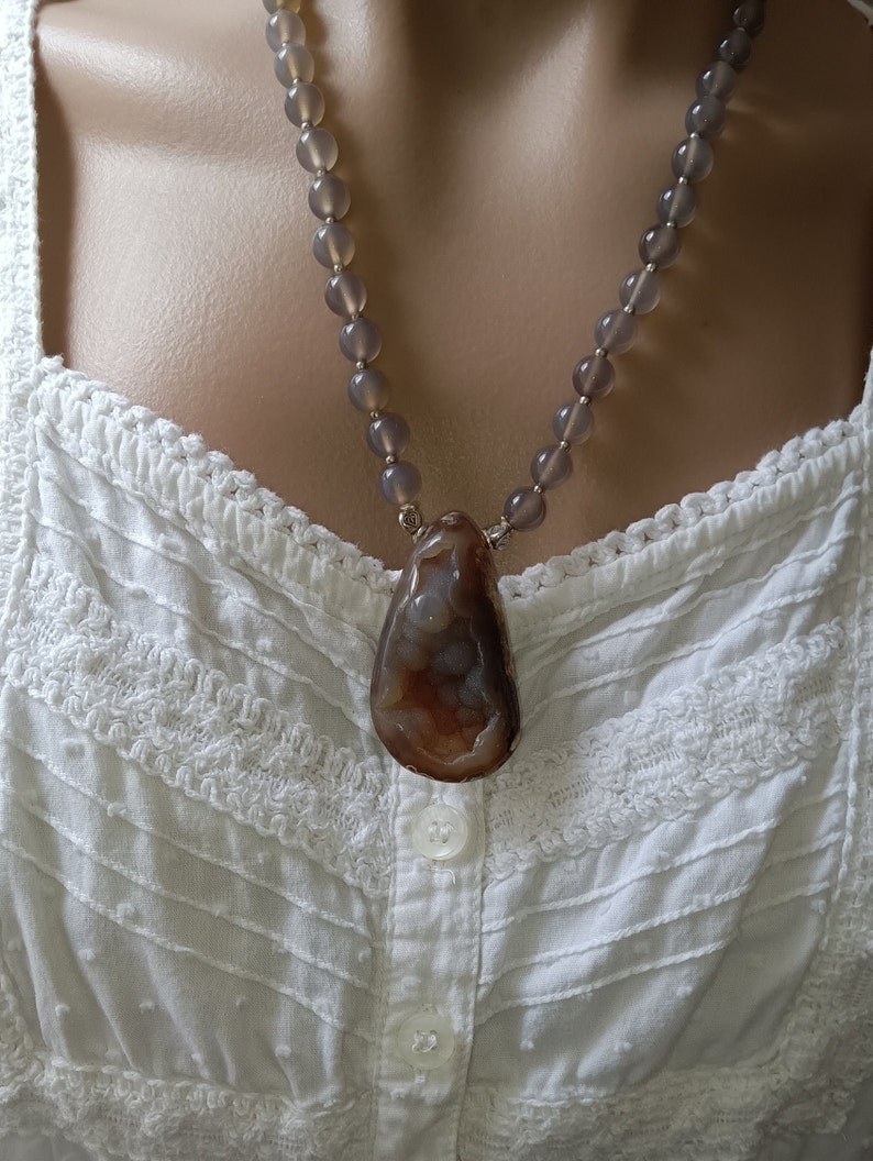 May include: A gray beaded necklace with a large brown agate pendant. The necklace is worn on a white lace top.