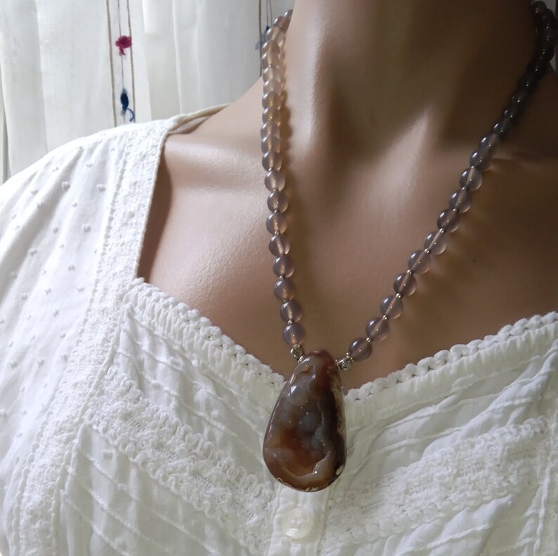May include: A long necklace made of light purple and gray beads with a large brown agate pendant.