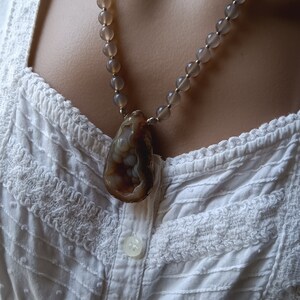 May include: A necklace with a large, brown agate pendant and gray beads. The necklace is worn on a white, lace-trimmed blouse.