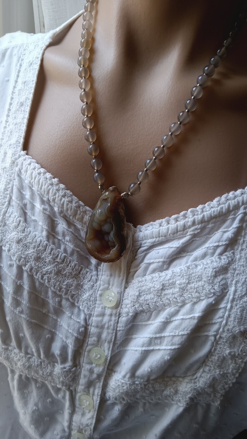 May include: A necklace with a large, brown agate pendant and gray beads. The necklace is worn on a white, lace-trimmed blouse.