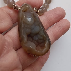May include: A brown agate pendant with a unique, intricate, and detailed pattern of white and gray agate crystals inside. The pendant is attached to a gray beaded necklace.