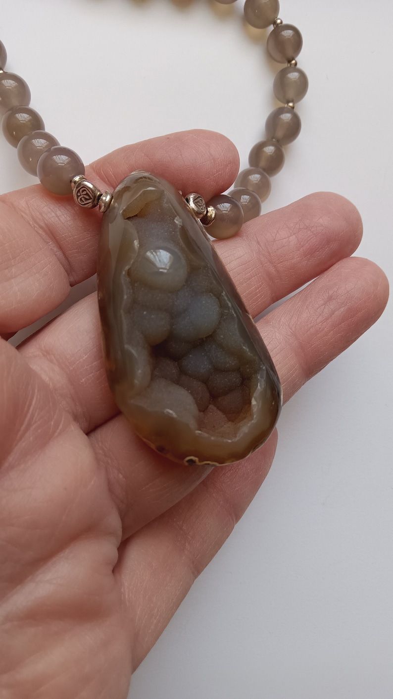 May include: A brown agate pendant with a unique, intricate, and detailed pattern of white and gray agate crystals inside. The pendant is attached to a gray beaded necklace.