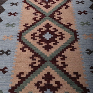 Könnte beinhalten: Ein gewebter Teppich mit einem sich wiederholenden geometrischen Muster in den Farben Hellblau, Beige, Dunkelkastanienbraun und Salbeigrün. Das Design besteht aus großen zentralen Rauten, die mit Dunkelkastanienbraun und Salbeigrün umrandet sind.