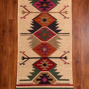 May include: A rectangular woven rug featuring a beige base with a repeating vertical pattern of diamonds and geometric shapes in shades of dark green, maroon, orange, black, and tan. The rug has fringed edges at the top and bottom ends.