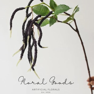 May include: An artificial plant stem featuring long, dark purple, textured catkin-like blooms with pale green tips and several green leaves on a brown branch. The text "Floral Goods ARTIFICIAL FLORALS Est. 2026" is printed at the bottom.
