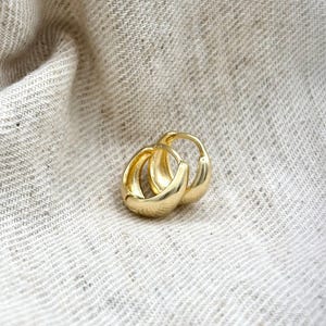 May include: A pair of gold-coloured hoop earrings. The earrings have a rounded, polished design. They are resting on a textured, off-white fabric.