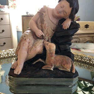 May include: A vintage figurine of a sleeping girl with a lamb. The girl is wearing a simple dress and has dark hair. The lamb is lying next to her and is looking up at her. The figurine is made of plaster and is painted in muted tones.