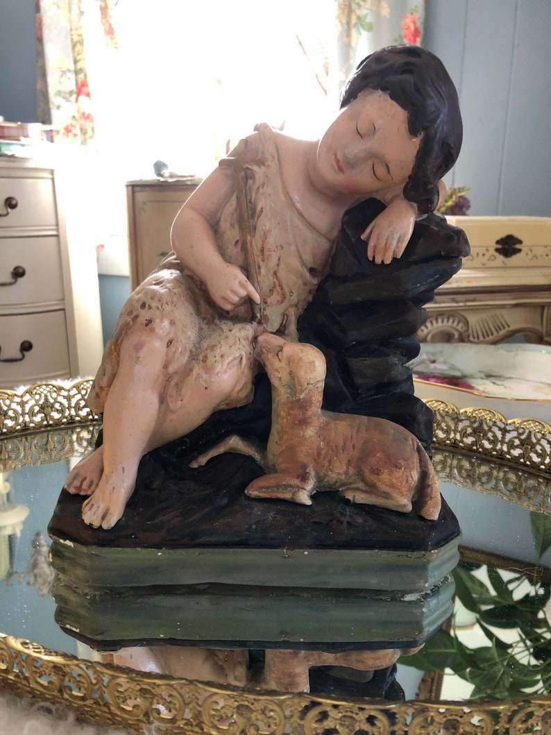 May include: A vintage figurine of a sleeping girl with a lamb. The girl is wearing a simple dress and has dark hair. The lamb is lying next to her and is looking up at her. The figurine is made of plaster and is painted in muted tones.