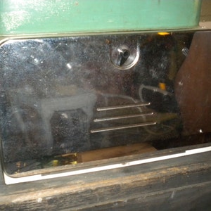 Op de afbeelding: Een vintage chromen broodtrommel met een witte rand en een metalen deur. De deur heeft een klein handvat en een reeks horizontale lijnen.