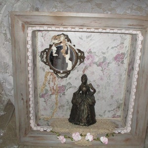 May include: A framed shadow box with a vintage mirror, a small figurine of a woman in a dress, and lace trim. The background is a floral patterned paper.