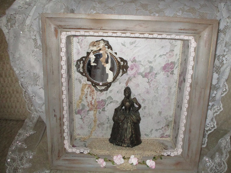 May include: A framed shadow box with a vintage mirror, a small figurine of a woman in a dress, and lace trim. The background is a floral patterned paper.