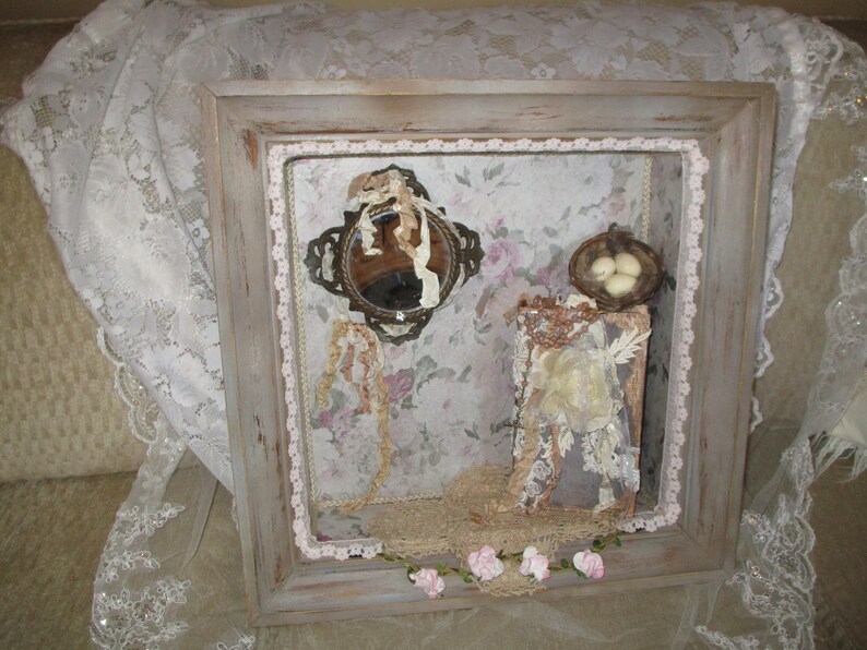 May include: A framed shadow box with a vintage mirror, a small bird's nest with three white eggs, and lace and floral accents. The box is painted in a distressed white and gray finish.
