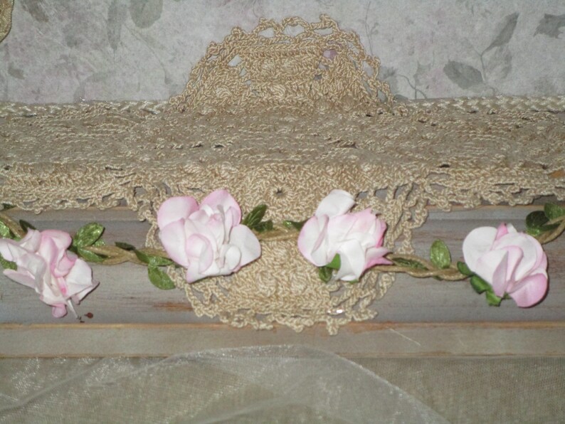May include: A close-up of a crocheted lace doily with a garland of pink fabric flowers. The doily is beige and the flowers are a light pink.