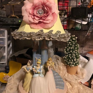 May include: A yellow lampshade with a pink flower and lace trim sits on a base with three Disney princesses: Cinderella, Snow White, and Aurora. The base is a light blue castle with a pink base.