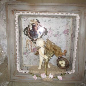 May include: A framed shadow box with a white porcelain cherub holding a gold horn, a small mirror, a bird's nest with eggs, and pink flowers.