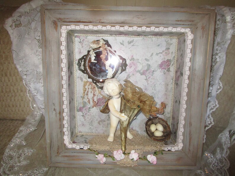 May include: A framed shadow box with a white porcelain cherub holding a gold horn, a small mirror, a bird's nest with eggs, and pink flowers.