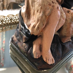 May include: A vintage plaster statue of a woman and child, painted in muted tones of brown, beige, and green. The statue is in a seated position, with the woman holding the child. The statue is on a base with a green and brown painted finish.