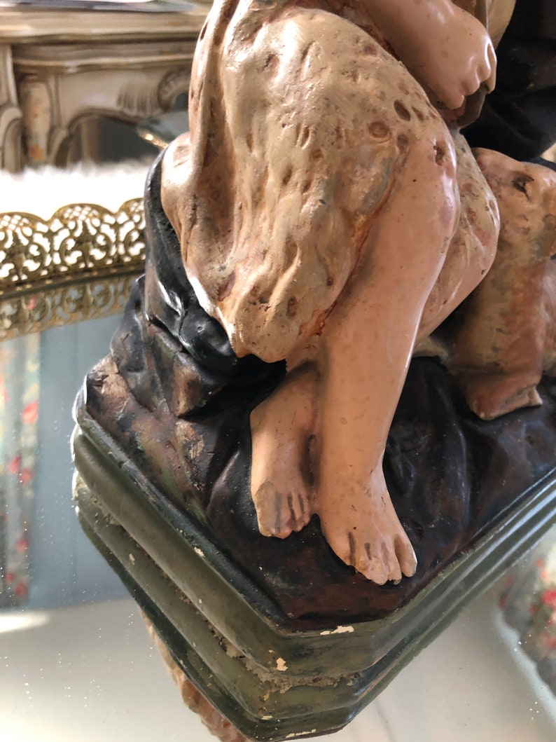May include: A vintage plaster statue of a woman and child, painted in muted tones of brown, beige, and green. The statue is in a seated position, with the woman holding the child. The statue is on a base with a green and brown painted finish.