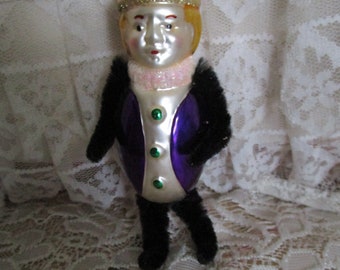 May include: A vintage Christmas ornament shaped like a regal figure. The ornament features a red and white hat, a face with blonde hair, and a purple and white vest. The figure has black fuzzy arms and legs, and a pink collar.