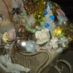 May include: A white sleigh filled with Christmas decorations, including pink roses, white roses, a small white angel, a bottle of perfume, and a variety of ornaments. The sleigh is decorated with gold glitter and white lace.