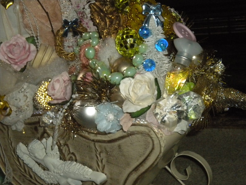 May include: A white sleigh filled with Christmas decorations, including pink roses, white roses, a small white angel, a bottle of perfume, and a variety of ornaments. The sleigh is decorated with gold glitter and white lace.