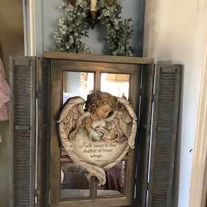 May include: A wooden window frame with shutters on either side. The window is a mirror and has a heart-shaped angel figurine with wings and a pink ribbon. The angel is holding its hands together in prayer. The ribbon has the text "I will trust in the shelter of Your wings. Psalm 61:4".