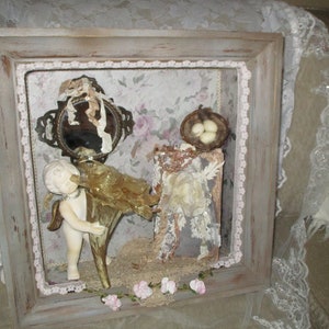 May include: A framed shadow box with a vintage aesthetic. It features a porcelain cherub figurine holding a golden trumpet, a small mirror with a decorative frame, a bird's nest with three white eggs, and a book with lace and beads. The box is decorated with lace and flowers.