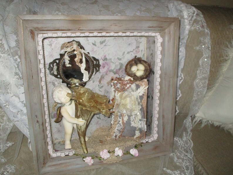 May include: A framed shadow box with a vintage aesthetic. It features a porcelain cherub figurine holding a golden trumpet, a small mirror with a decorative frame, a bird's nest with three white eggs, and a book with lace and beads. The box is decorated with lace and flowers.