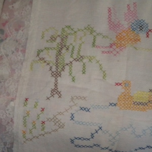 May include: White fabric with colorful cross-stitch embroidery of a tree, a bird, and a duck. The bird is pink, orange, and blue. The duck is orange and yellow. The tree is green and brown.