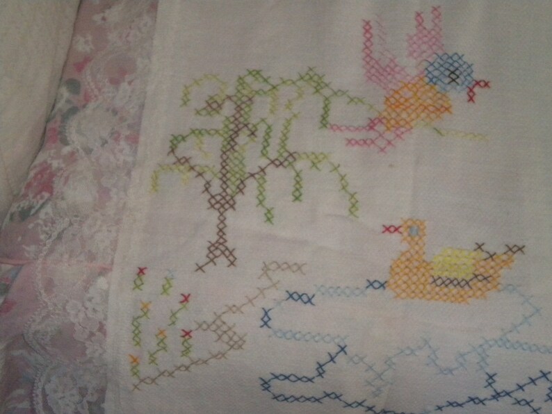 May include: White fabric with colorful cross-stitch embroidery of a tree, a bird, and a duck. The bird is pink, orange, and blue. The duck is orange and yellow. The tree is green and brown.